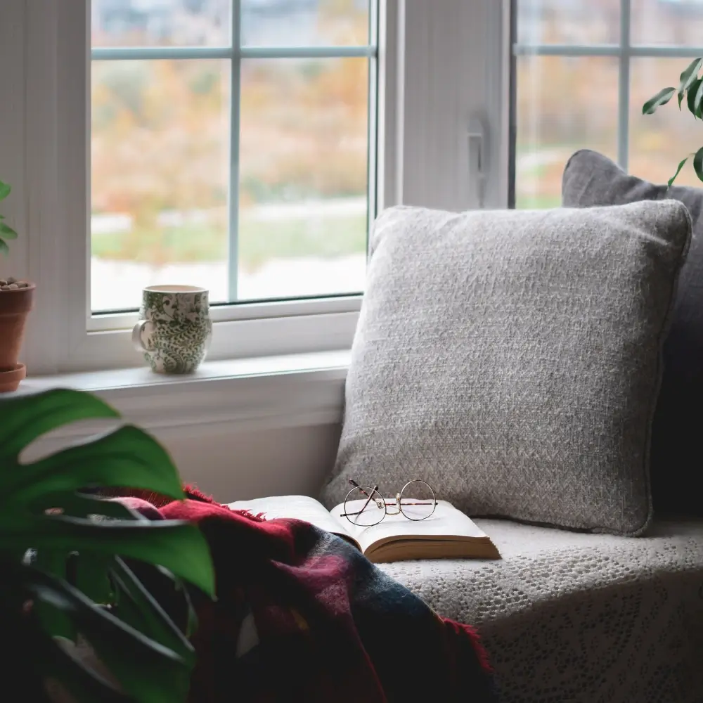 comfy nook sunlight coming in coffee cup on the window ledge open book with glasses on it greenery in the foreground perfect for a relaxing morning reading
